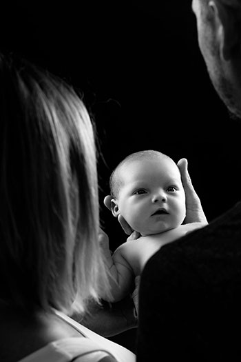 Photo d'un bébé nouveau-né de deux semaines en noir et blanc, le bébé regarde les yeux de son père qui le soutien dans ses mains.