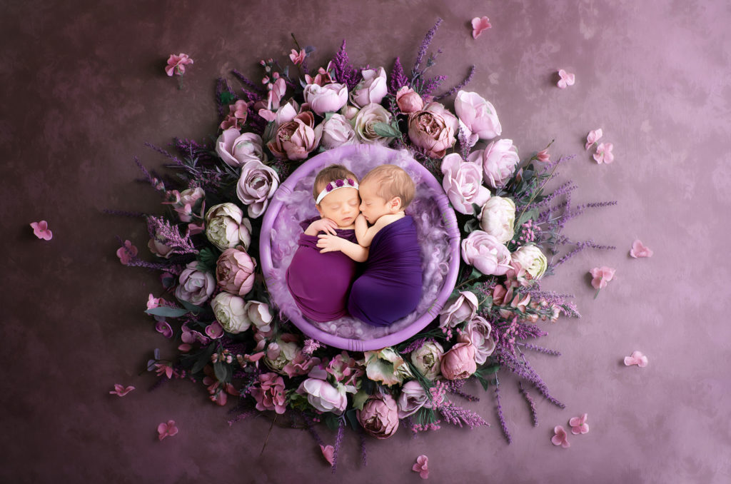 Photographie de bébés jumeaux endormis, entourés de roses et de fleurs violettes.
